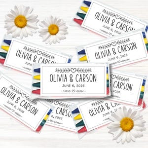 以下が含まれることがあります： OLIVIA & CARSONおよびJUNE 6, 2026という文字と、ハートや葉の枝の黒い線画が描かれた白いラベルが付いた、多色のクレヨンが入った透明なプラスチックパックが複数あり、白いデイジーの花と一緒に配置されています。