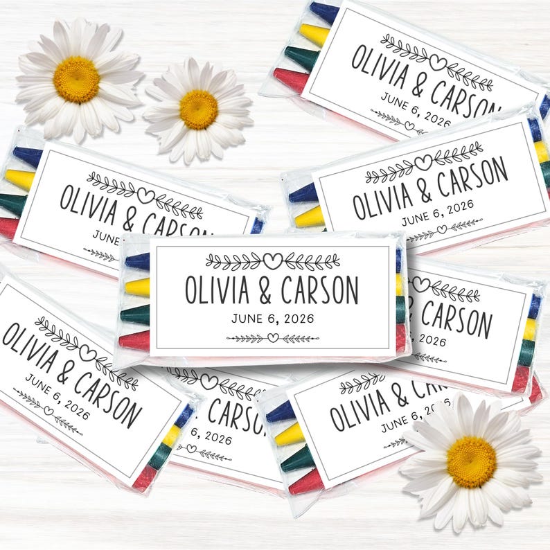 以下が含まれることがあります： OLIVIA & CARSONおよびJUNE 6, 2026という文字と、ハートや葉の枝の黒い線画が描かれた白いラベルが付いた、多色のクレヨンが入った透明なプラスチックパックが複数あり、白いデイジーの花と一緒に配置されています。
