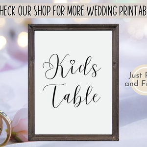 May include: Printable sign with a brown frame that says "Kids Table" in a cursive font with a small heart symbol above the word "Kids". The sign is on a white surface with a pink floral pattern.