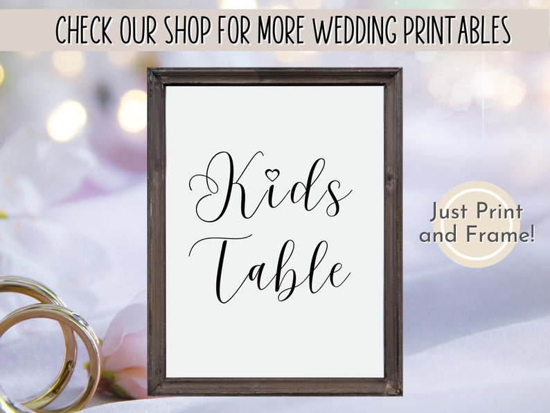May include: Printable sign with a brown frame that says "Kids Table" in a cursive font with a small heart symbol above the word "Kids". The sign is on a white surface with a pink floral pattern.