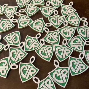 Puede incluir: Una colección de colgantes verdes y blancos. Cada colgante tiene forma de escudo con un lazo en la parte superior y presenta las letras "CTR" en una fuente estilizada. Los colgantes están dispersos sobre una superficie marrón oscuro.