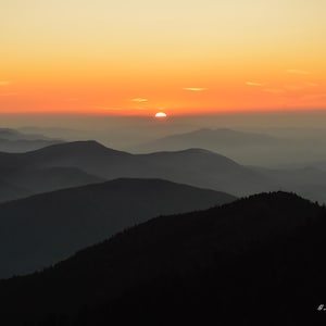 May include: A landscape photograph of a mountain range at sunset. The sky is a gradient of orange and yellow, with the sun partially visible. The mountains are silhouetted in shades of grey and black.