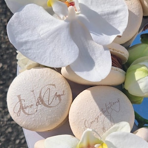 May include: A close-up of decorated macarons with white orchid flowers. Some macarons have the names "Jalsena & Amit" and "Congratulations" written on them in gold script. The macarons are light beige with a chocolate filling.