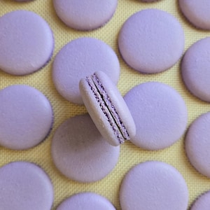 May include: A close-up of a baking sheet filled with lavender-colored macarons. The macarons are arranged in a grid pattern, and one macaron is slightly offset from the others.