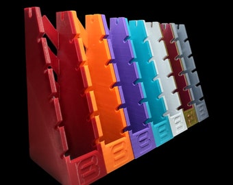 May include: A set of seven colorful plastic display stands, each with a unique color, including red, orange, purple, blue, white, maroon, and gray. The stands are angled and have a series of slots for displaying items. Each stand has the letter "B" embossed on the bottom.