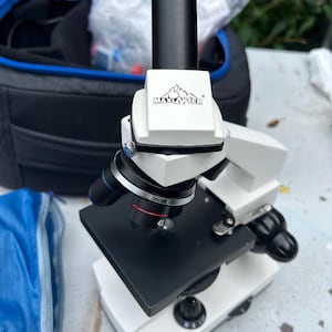 Op de afbeelding: Een wit en zwart Maxlapter samengestelde microscoop met een draaibare revolverkop met meerdere objectieflenzen, waaronder een gemarkeerd met 4X. Het apparaat bevat een zwarte tafel, een verticale oculairbuis en instelknoppen voor focus.