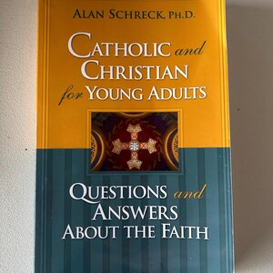 May include: A hardcover book titled "Catholic and Christian for Young Adults" by Alan Schreck, Ph.D. The cover is split horizontally, with an orange top section and a teal bottom section. The book includes the text "Questions and Answers About the Faith."