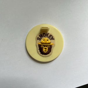 May include: A yellow plastic round button with a brown and yellow embroidered Smokey Bear logo. The button has a small hole at the top.