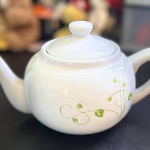 Könnte beinhalten: Weiße Teekanne aus Keramik mit grünem Blumendesign. Die Teekanne hat einen Deckel und einen Ausguss.