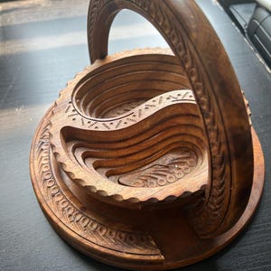 May include: A wooden serving bowl with a carved handle. The bowl is made of dark brown wood and has a circular shape. The bowl is divided into sections, and the handle is attached to the top of the bowl.