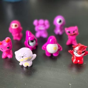 May include: A collection of ten small, colorful plastic figurines. The figurines are in various shapes and colors, including pink, purple, and red. They have cartoon-like features and are designed to look like characters from a children's cartoon.