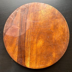 May include: A round wooden cutting board with a natural brown finish. The board has a smooth surface and is perfect for chopping vegetables or serving cheese.