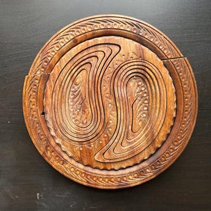 May include: A round wooden serving tray with intricate carvings. The tray is made of dark brown wood and has a circular design with a center that is divided into two sections. The tray is perfect for serving snacks, appetizers, or desserts.