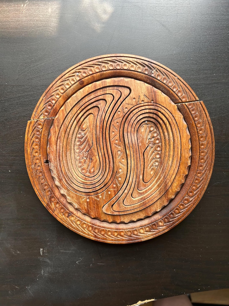 May include: A round wooden serving tray with intricate carvings. The tray is made of dark brown wood and has a circular design with a center that is divided into two sections. The tray is perfect for serving snacks, appetizers, or desserts.