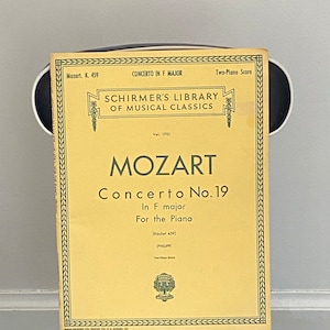 May include: A yellow sheet music cover for Mozart's Piano Concerto No. 19 in F major, from Schirmer's Library of Musical Classics. The cover features a decorative border and the text "Mozart Concerto No. 19 In F major For the Piano [Köchel 459] PHILIPP Two-Piano Score".