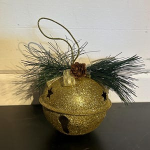 Peut inclure: Une cloche de Noël dorée et scintillante avec des découpes en forme d'étoiles. La cloche est ornée de fausses aiguilles de pin, d'une petite pomme de pin et d'un ruban doré. Une boucle en fil d'or est attachée pour l'accrochage.