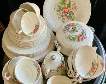 May include: A collection of cream-colored vintage china with embossed floral patterns. The set includes cups, saucers, bowls, a serving bowl, a covered dish, a creamer, and a sugar bowl. Each piece features a colorful floral design.