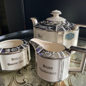 May include: A white teapot, creamer and sugar bowl with silver trim and black floral designs. Each piece has the words "Happy Anniversary" written on it. The teapot has a blue label that reads "Sadler Fine English Teapots Co."