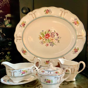 May include: A vintage porcelain serving set featuring a floral pattern. The set comprises a large oval platter, a creamer, a sugar bowl with a lid, and a gravy boat with a matching saucer. The dishes are decorated with a delicate floral design in shades of pink, yellow, and blue, accented with gold trim.