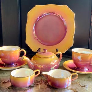 Puede incluir: Un juego de té vintage con un acabado iridiscente. El juego incluye un plato grande, una tetera, una crema, un azucarero y dos tazas de té con platillos. Los colores van del amarillo al naranja y al rosa.