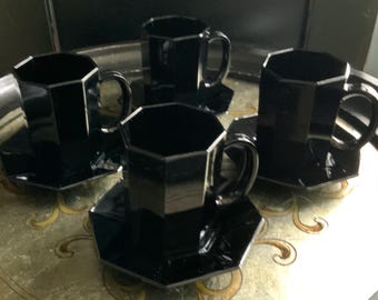 May include: Four black glass teacups and saucers are arranged on a decorative tray. The cups and saucers have a geometric, octagonal design. The tray has a gold and cream colored floral pattern.