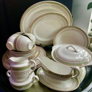 May include: A collection of cream-coloured ceramic dinnerware featuring a repeating green and brown geometric border pattern with gold trim. The set includes oval platters, a lidded tureen, a gravy boat with a stand, a creamer, and stacked plates.