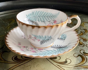 Puede incluir: Juego de taza y platillo blancos con borde dorado y diseño de helechos y flores en tonos azules, rosas y negros. La taza tiene un diseño acanalado. Juego de té probablemente vintage o antiguo.