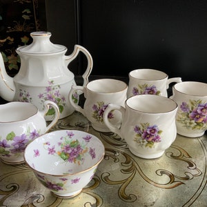 Puede incluir: Un juego de té blanco con un patrón floral de violetas moradas y verdes. El juego incluye una tetera, una jarra de crema, un azucarero y cuatro tazas de té.
