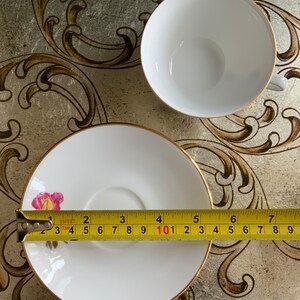 以下が含まれることがあります： 金色の縁取りが施された白いティーカップとソーサーのセット。ソーサーにはピンクのバラのデザインが施されています。ティーカップとソーサーの直径は12.7cmです。