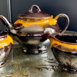 Peut inclure: Un service à thé en céramique brun et jaune avec une théière, un pot à crème et un sucrier. L'ensemble présente un motif floral et une finition brillante.