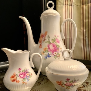 Puede incluir: Un juego de té de porcelana blanca con un diseño floral. El juego incluye una tetera, una jarra de crema y un azucarero. Las flores son rosas, naranjas y azules.