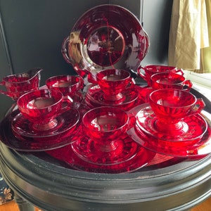 Puede incluir: Un juego completo de cristalería rojo rubí, que incluye tazas, platillos, platos, una fuente para servir y una jarra para crema. La cristalería está dispuesta sobre una mesa redonda y negra. El juego está hecho de vidrio transparente texturizado.