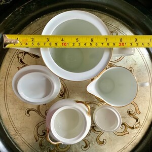 以下が含まれることがあります： 金色の縁取りが施された白い磁器製のティーセット。セットにはティーポット、クリーマー、シュガーボウル、蓋付きカップが含まれています。セットは金色のデザインが施された装飾的なトレイの上に置かれています。