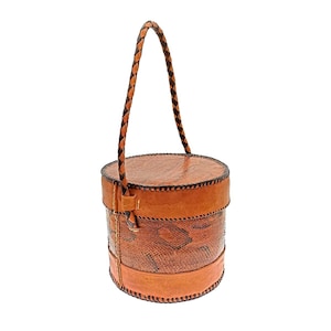 May include: A cylindrical, brown leather handbag with a braided handle. The bag features a snakeskin-like pattern on the body, with stitched detailing and a solid leather top and bottom. The handle is a dark brown, braided design.