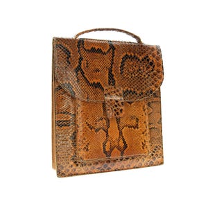 Peut inclure: Une mallette en peau de serpent marron et noire avec une poignée supérieure et une fermeture à rabat. La mallette a une forme rectangulaire avec une poche avant et une boucle. Le motif peau de serpent est visible sur tout le sac.