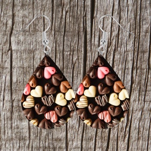 Może przedstawiać: Kolczyki w kształcie łezki z motywem czekoladowych cukierków. Kolczyki mają ciemnobrązowe tło z cukierkami w kształcie serca w odcieniach brązu, różu i bieli. Kolczyki mają srebrne haczyki.