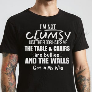 May include: Black crew neck t-shirt with white text that reads "I'M NOT CLUMSY JUST THE FLOOR HATES ME THE TABLE & CHAIRS are bullies AND THE WALLS Get in My Way". The shirt is a casual wear item.