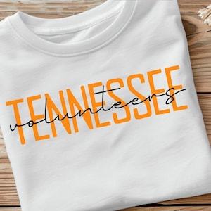 Puede incluir: Camiseta blanca con la frase "Tennessee Volunteers" impresa en letras naranja y escritura cursiva negra. La camiseta está doblada y sobre una superficie de madera.