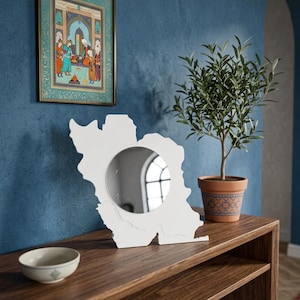 May include: A white mirror in the shape of the country of Iran sits on a wooden table. A small potted olive tree and a framed painting are also on the table. A small ceramic bowl is on the table.
