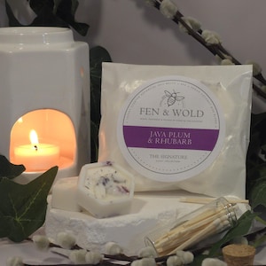 May include: A white ceramic wax warmer with a lit tea light inside. Next to it is a white wax melt in a hexagonal shape with a floral design. A small glass jar filled with wooden matches is also visible. The wax melt is labelled "Java Plum & Rhubarb" and "The Signature Scent Collection" by Fen & Wold.