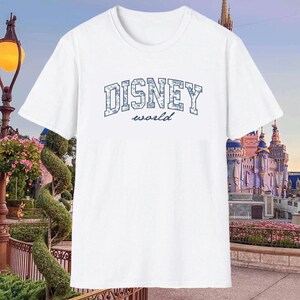 K&ouml;nnte beinhalten: Wei&szlig;es T-Shirt mit dem Text "DISNEY world" in Blau. Das Wort "DISNEY" ist in Blockbuchstaben geschrieben. Das Shirt hat einen Rundhalsausschnitt und kurze &Auml;rmel. Der Hintergrund zeigt einen Themenpark mit einem Schloss.