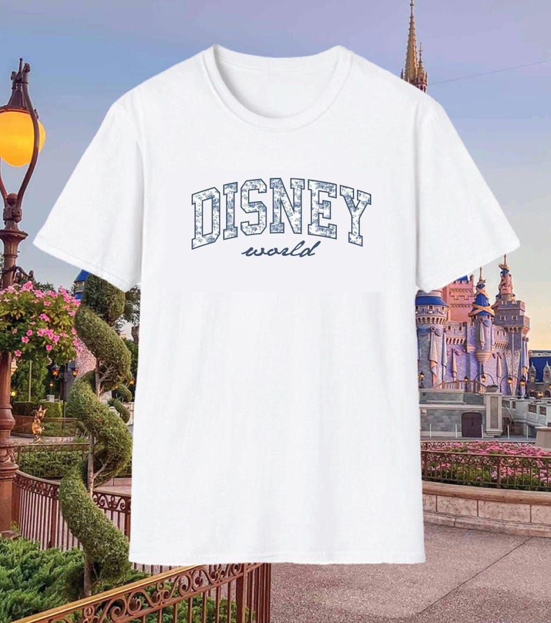 K&ouml;nnte beinhalten: Wei&szlig;es T-Shirt mit dem Text "DISNEY world" in Blau. Das Wort "DISNEY" ist in Blockbuchstaben geschrieben. Das Shirt hat einen Rundhalsausschnitt und kurze &Auml;rmel. Der Hintergrund zeigt einen Themenpark mit einem Schloss.