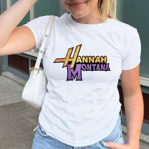 May include: White short-sleeved t-shirt featuring the text "Hannah Montana" in a colorful, stylized design. The design includes gold, purple, and black lettering. The shirt is made of a soft, comfortable material.