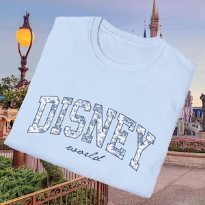 K&ouml;nnte beinhalten: Hellblaues T-Shirt mit dem Aufdruck "DISNEY world" in einem floralen Design. Das Wort "DISNEY" ist in einer fetten, bogenf&ouml;rmigen Schriftart gehalten, w&auml;hrend "world" in einer kleineren, kursiven Schriftart steht. Das Shirt hat einen Rundhalsausschnitt.