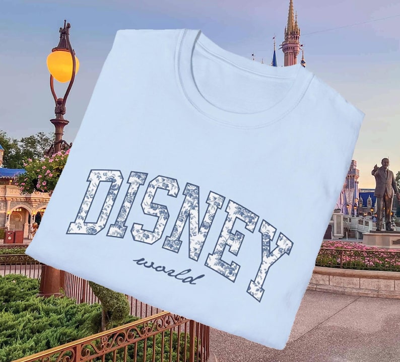 K&ouml;nnte beinhalten: Hellblaues T-Shirt mit dem Aufdruck "DISNEY world" in einem floralen Design. Das Wort "DISNEY" ist in einer fetten, bogenf&ouml;rmigen Schriftart gehalten, w&auml;hrend "world" in einer kleineren, kursiven Schriftart steht. Das Shirt hat einen Rundhalsausschnitt.