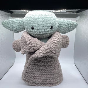 May include: A crocheted plush toy resembling a popular fictional character, wearing a brown crocheted blanket. The toy has large, green ears and black eyes.