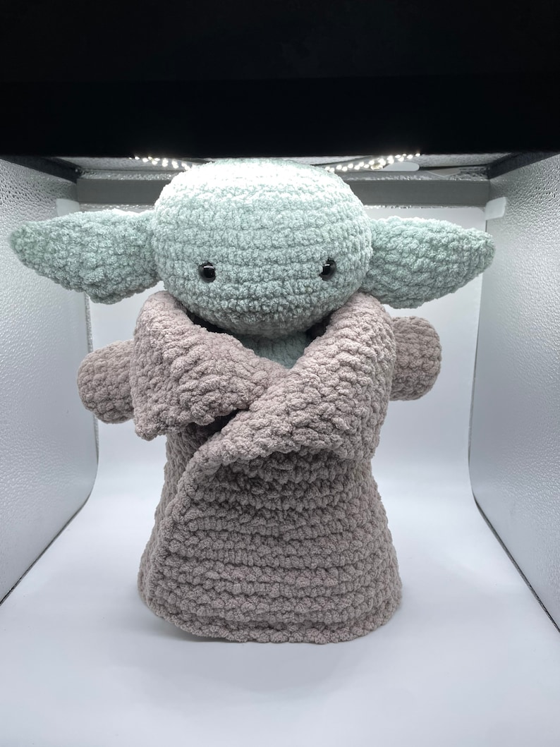 May include: A crocheted plush toy resembling a popular fictional character, wearing a brown crocheted blanket. The toy has large, green ears and black eyes.