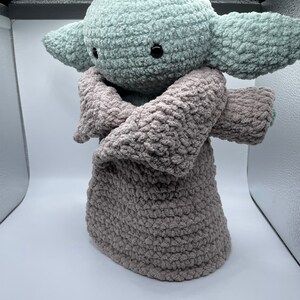 May include: A crocheted plush toy of a green character with large ears, wearing a brown crocheted robe. The toy is standing on a white surface.