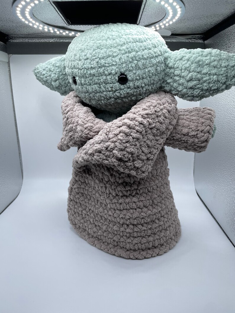 May include: A crocheted plush toy of a green character with large ears, wearing a brown crocheted robe. The toy is standing on a white surface.