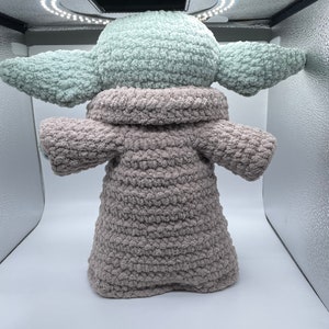 May include: A crocheted plush toy resembling a character from the Star Wars franchise. The toy is wearing a brown hooded robe and has a green head and ears.